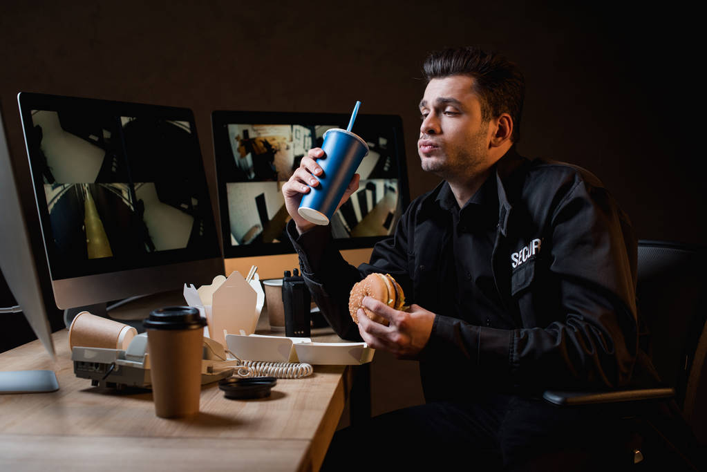 Strażnik jedzenie burger, trzymając kubek papierowy i patrząc na monitor komputera  - Zdjęcie, obraz