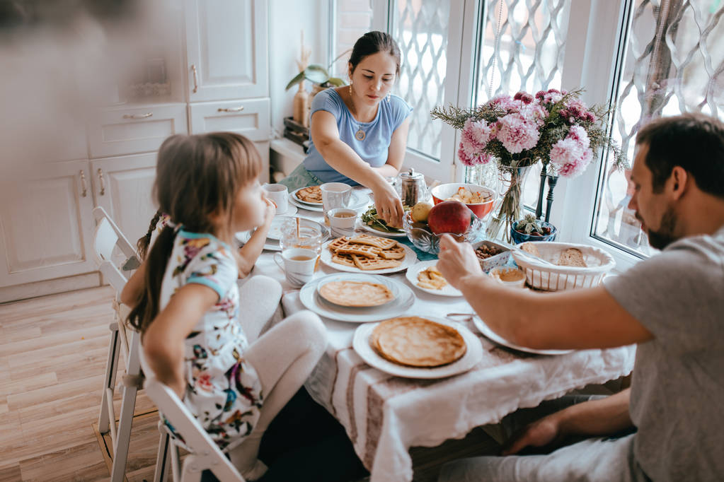 Family breakfast at home in the nice cozy kitchen. Mother, father and their two daughters eating pancakes - Photo, Image