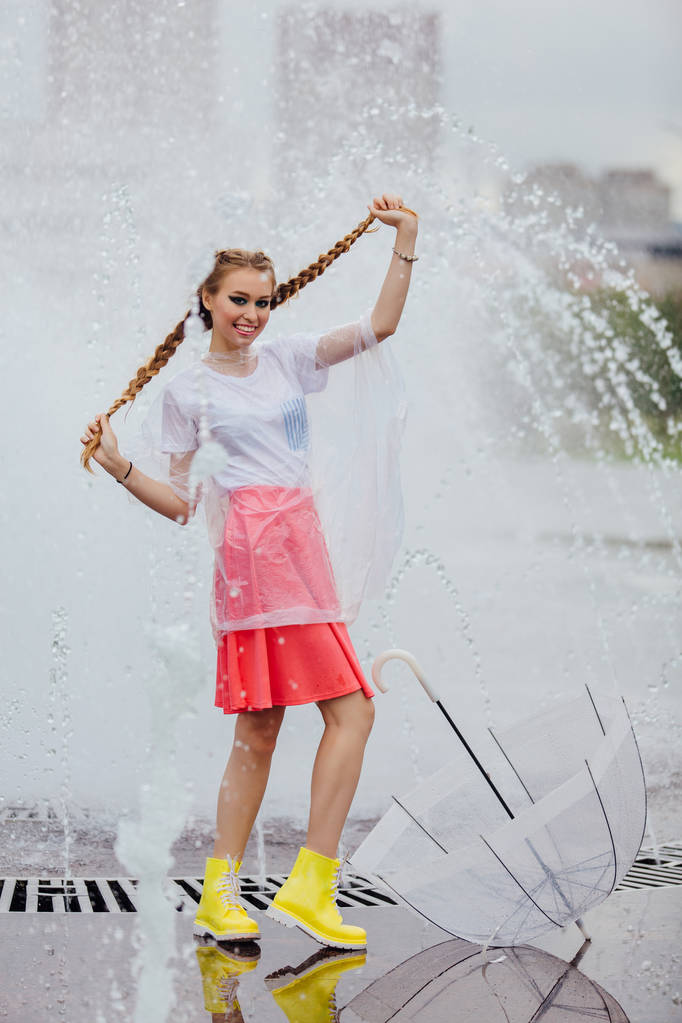 Mooi meisje met twee vlechten in gele laarzen en met transparante paraplu staat in de buurt van fontein. Regenachtige dag in de stad. - Foto, afbeelding