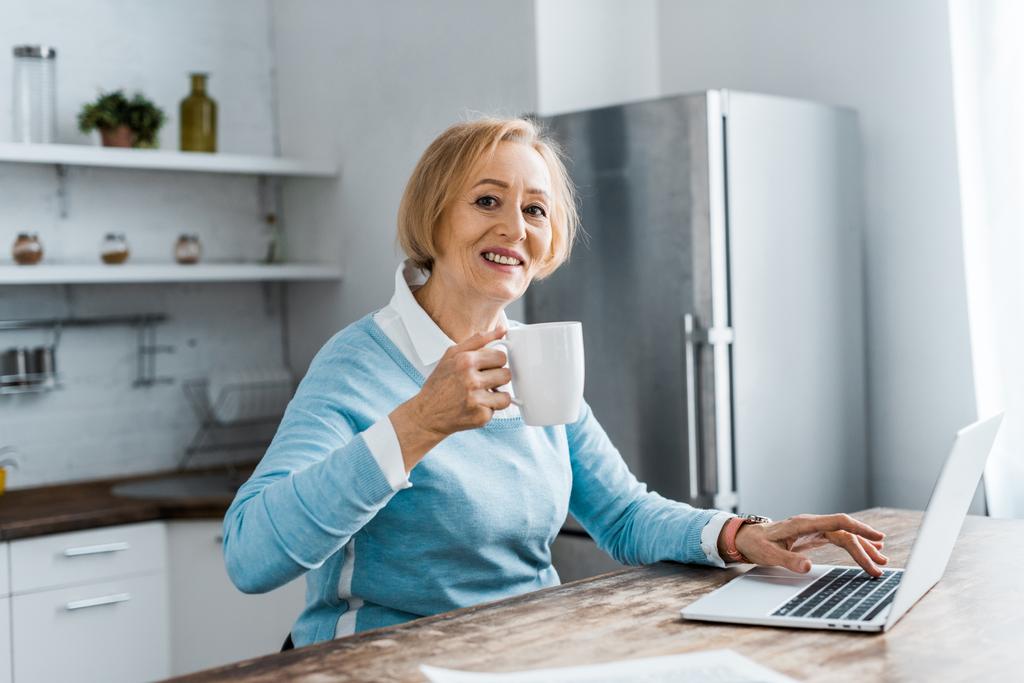 glimlachend senior vrouw zittend aan tafel, camera kijken, koffie te drinken en het gebruik van laptop in keuken - Foto, afbeelding