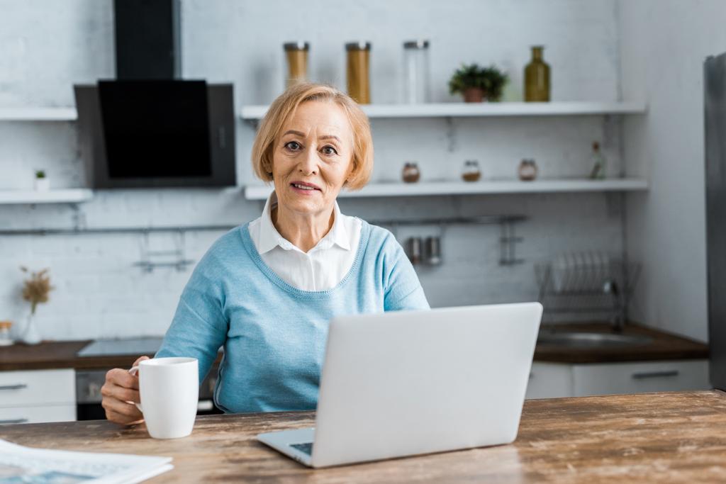 Senior vrouw zittend aan tafel met een kopje koffie, camera kijken en met behulp van laptop in de keuken - Foto, afbeelding