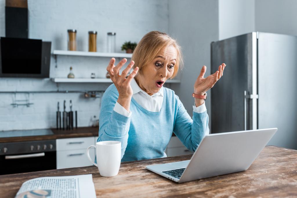 mulher sênior surpreso gesticular com as mãos, usando laptop e ter bate-papo de vídeo na cozinha
 - Foto, Imagem