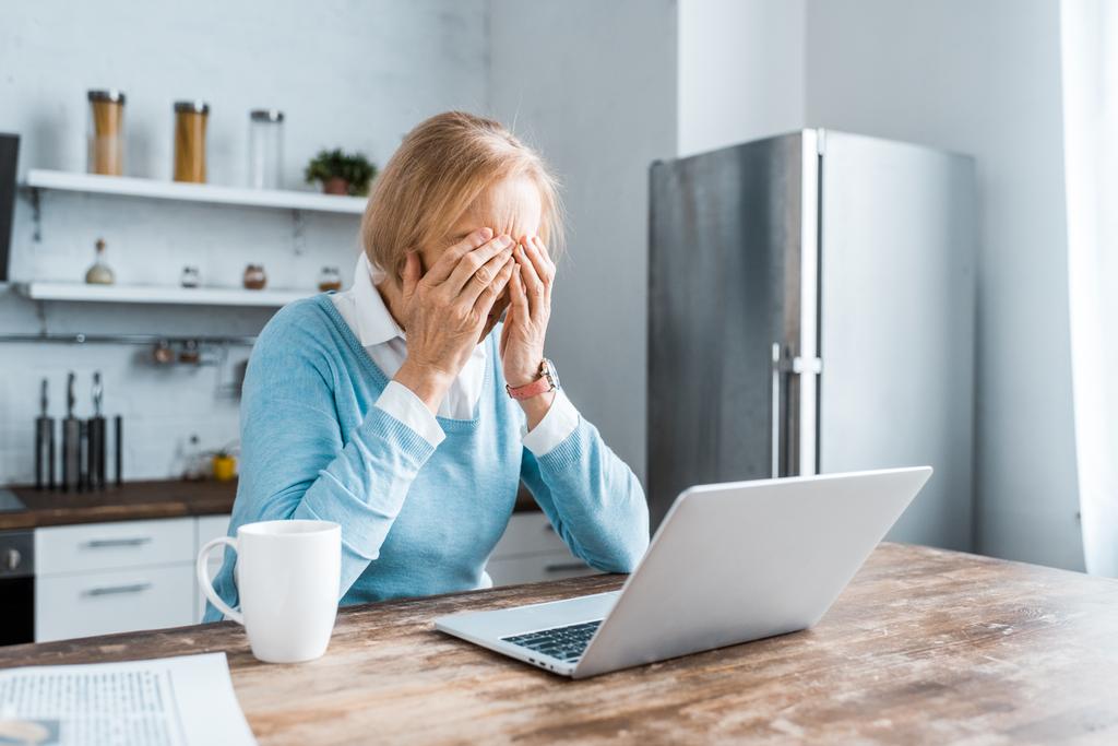 стресс старшая женщина сидит за столом и покрывает лицо во время видеозвонка на ноутбук на кухне
 - Фото, изображение