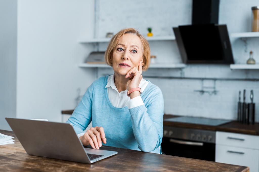 donna anziana pensierosa seduta a tavola con computer portatile e mento protettivo in cucina
 - Foto, immagini