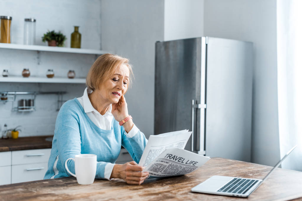 старша жінка сидить за столом з чашкою кави і ноутбуком під час читання газети на кухні
 - Фото, зображення