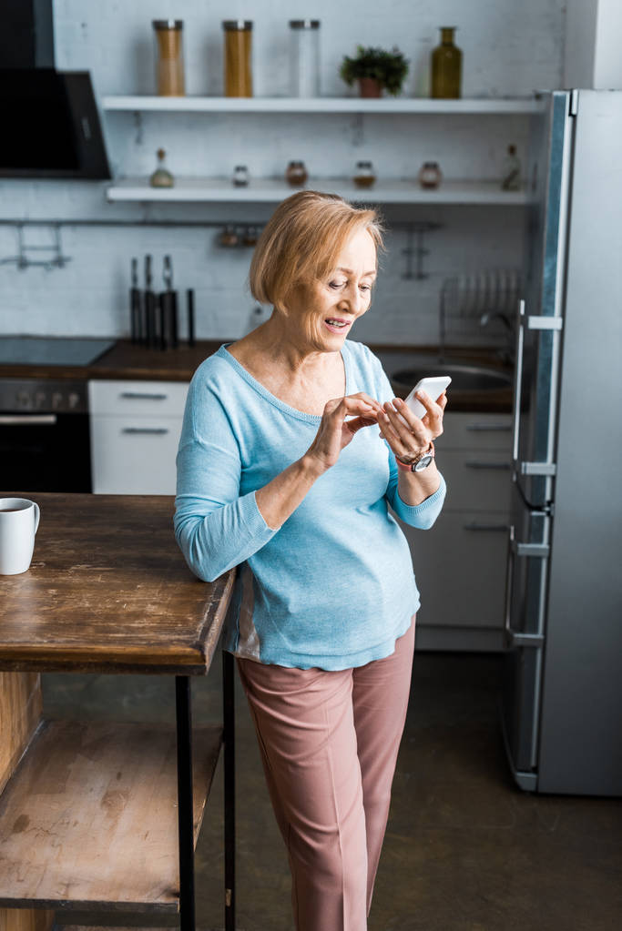 улыбающаяся пожилая женщина с помощью смартфона на кухне
  - Фото, изображение