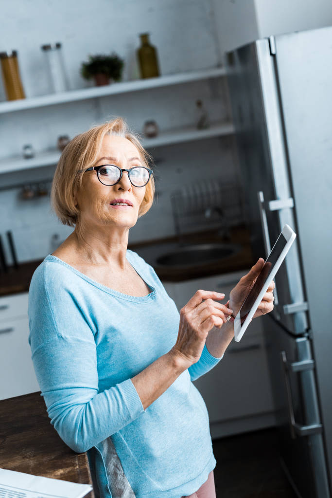 senior femme en lunettes regardant l'appareil photo et en utilisant une tablette numérique à la maison
 - Photo, image