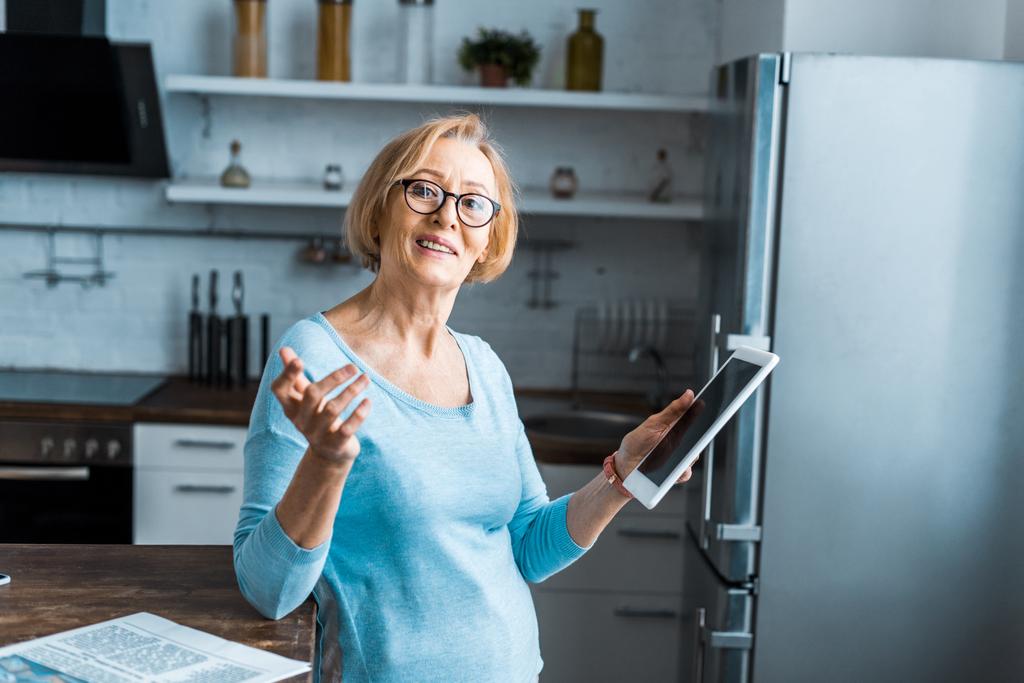 hymyilevä vanhempi nainen lasit käyttäen digitaalista tablettia ja eleitä käsin keittiössä
 - Valokuva, kuva