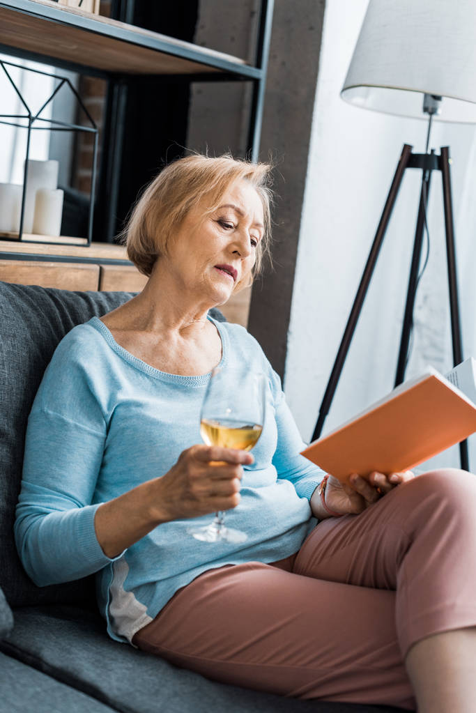 ανώτερος γυναίκα κάθεται στον καναπέ, κρατώντας το ποτήρι κρασί και διαβάζοντας το βιβλίο στο σπίτι - Φωτογραφία, εικόνα