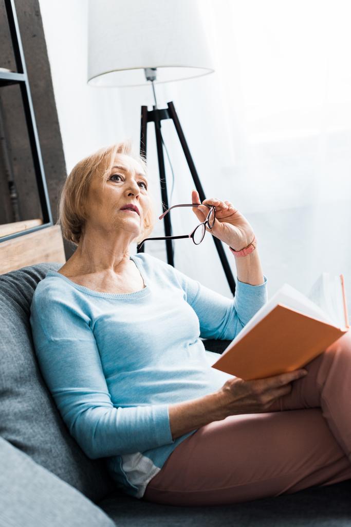 zamyšlený starší žena sedí na gauči, drží brýle a koukal při čtení knihy doma  - Fotografie, Obrázek