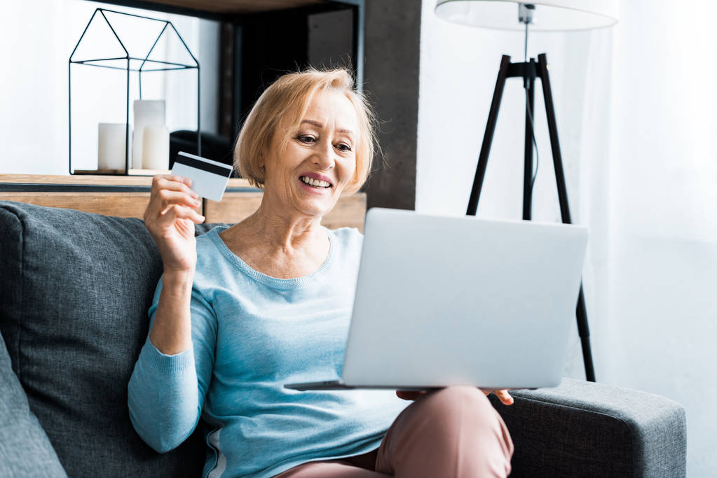 幸せな先輩女座っている、クレジット カードを保持していると家庭でオンライン ショッピングをしながらノート パソコンを使用して - 写真・画像