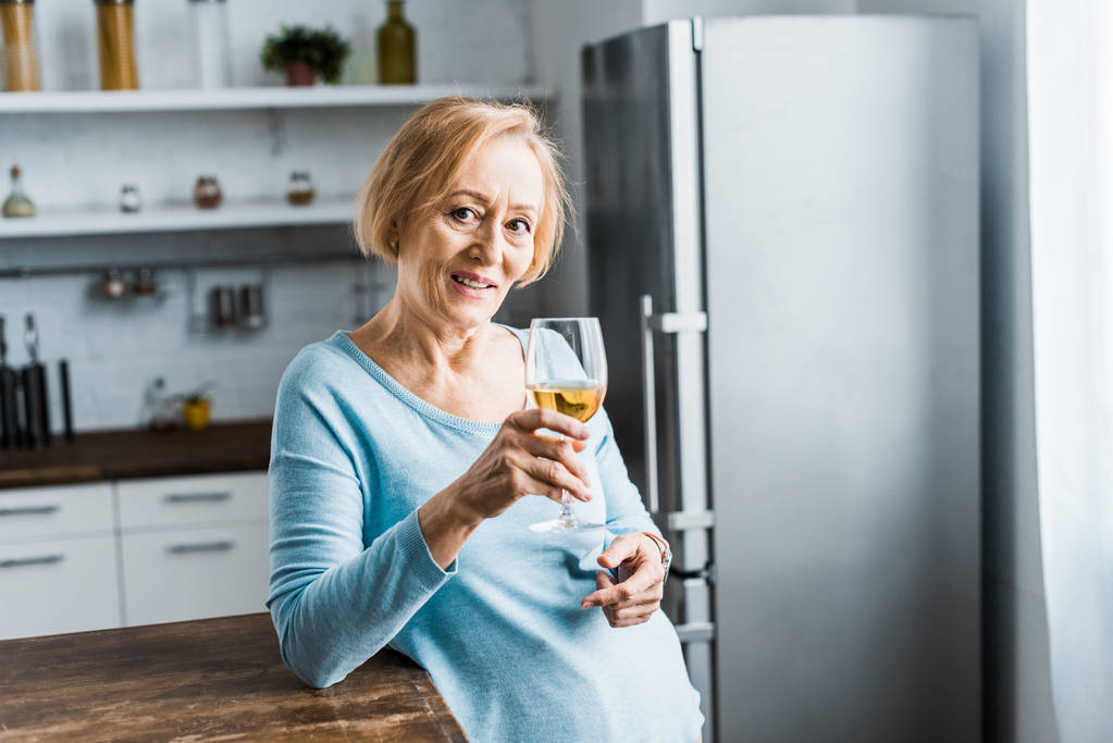 uśmiechnięta kobieta starszy patrząc na kamery i trzymając kieliszek do wina w kuchni z miejsca kopii - Zdjęcie, obraz