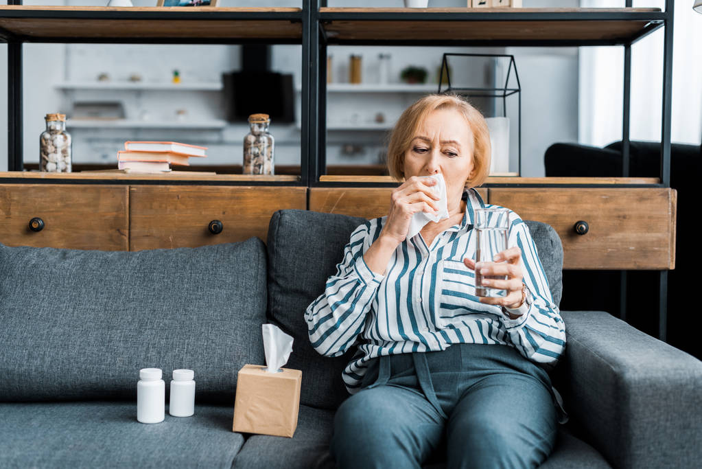 femme âgée malade avec nez qui coule assis sur le canapé avec une boîte en tissu et tenant un verre d'eau dans le salon
 - Photo, image