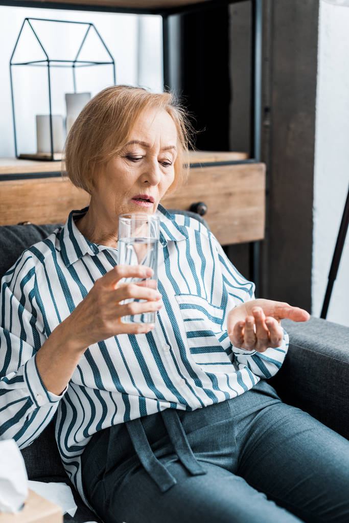 хвора старша жінка сидить і тримає ліки зі склянкою води вдома
 - Фото, зображення