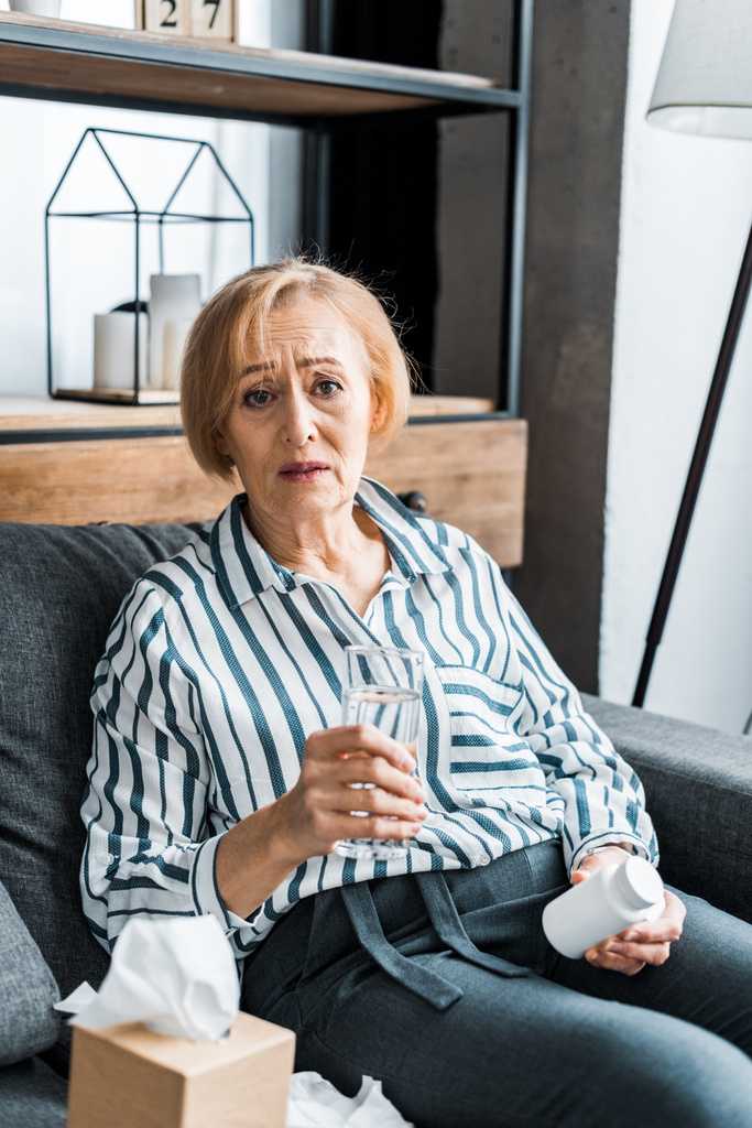 sick senior woman looking at camera and holding bottle of medication with glass of water  - Photo, Image