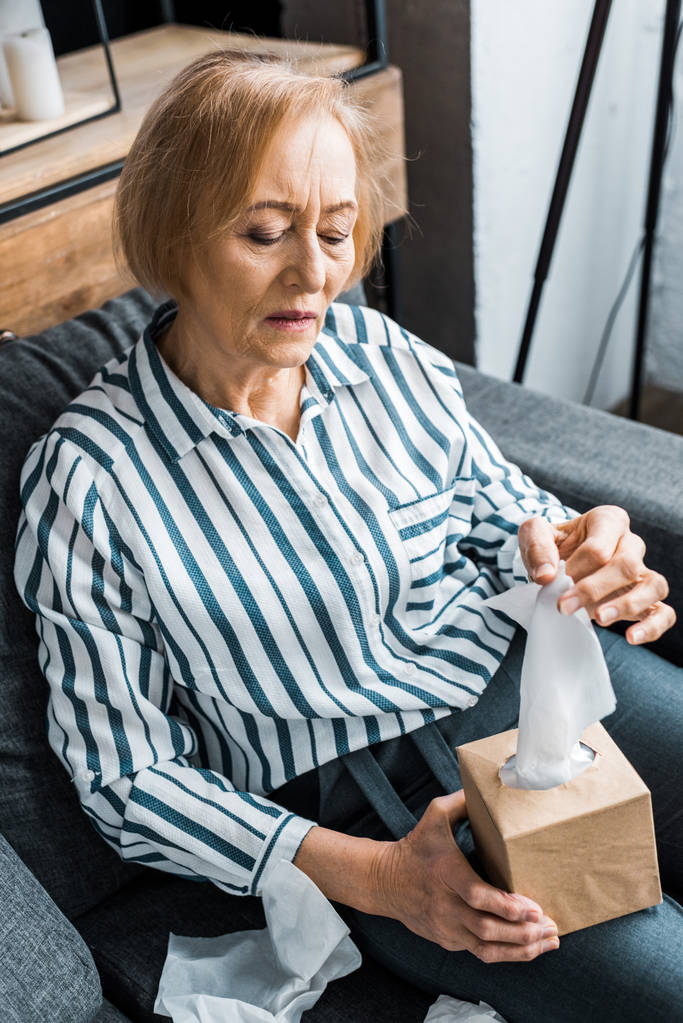 хвора старша жінка, яка страждає від застуди і досягає тканини, сидячи вдома
 - Фото, зображення