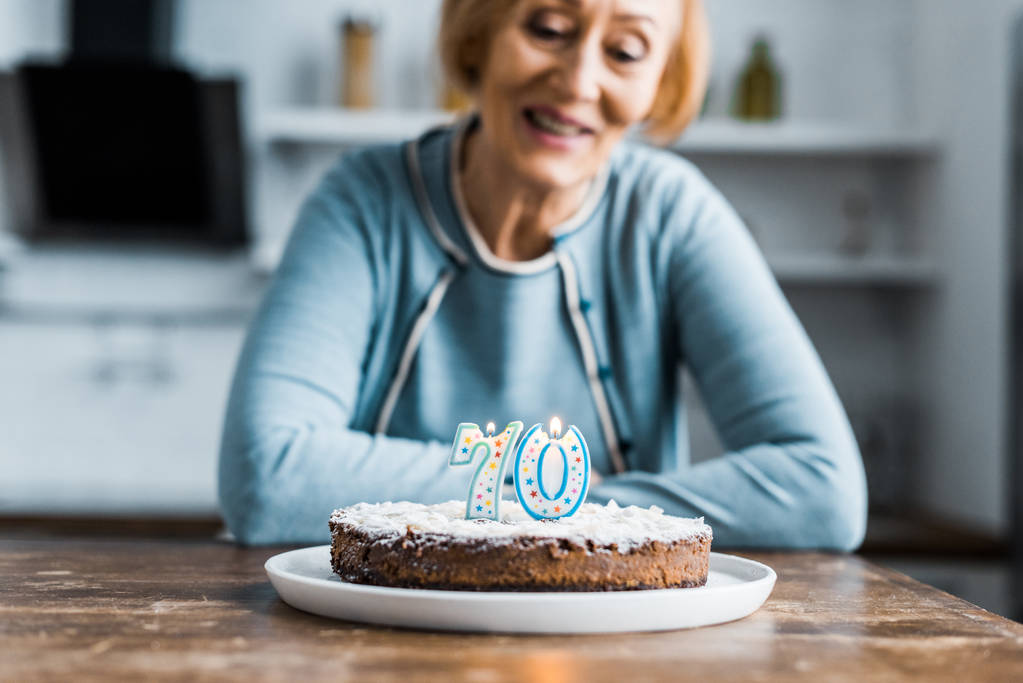 selective focus of cake with '70' sign on top and smiling senior woman on background during birthday celebration - Photo, Image