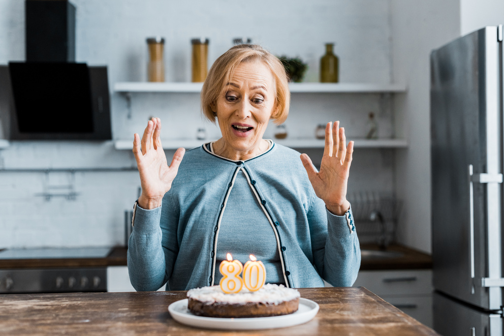 ενθουσιασμένος ανώτερων γυναίκα κάθεται και ψάχνει στο κέικ με το σύμβολο «80» στην κορυφή κατά τη γιορτή των γενεθλίων - Φωτογραφία, εικόνα