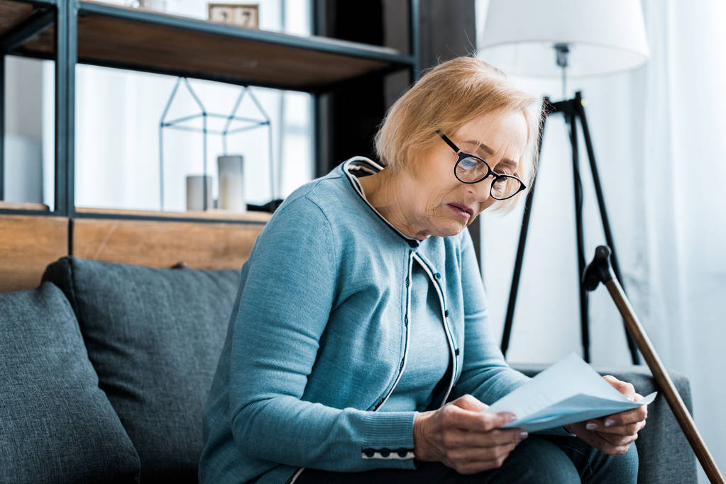 Senior kobietę w okularach, siedząc na kanapie i przytrzymując formularz podatkowy w domu - Zdjęcie, obraz