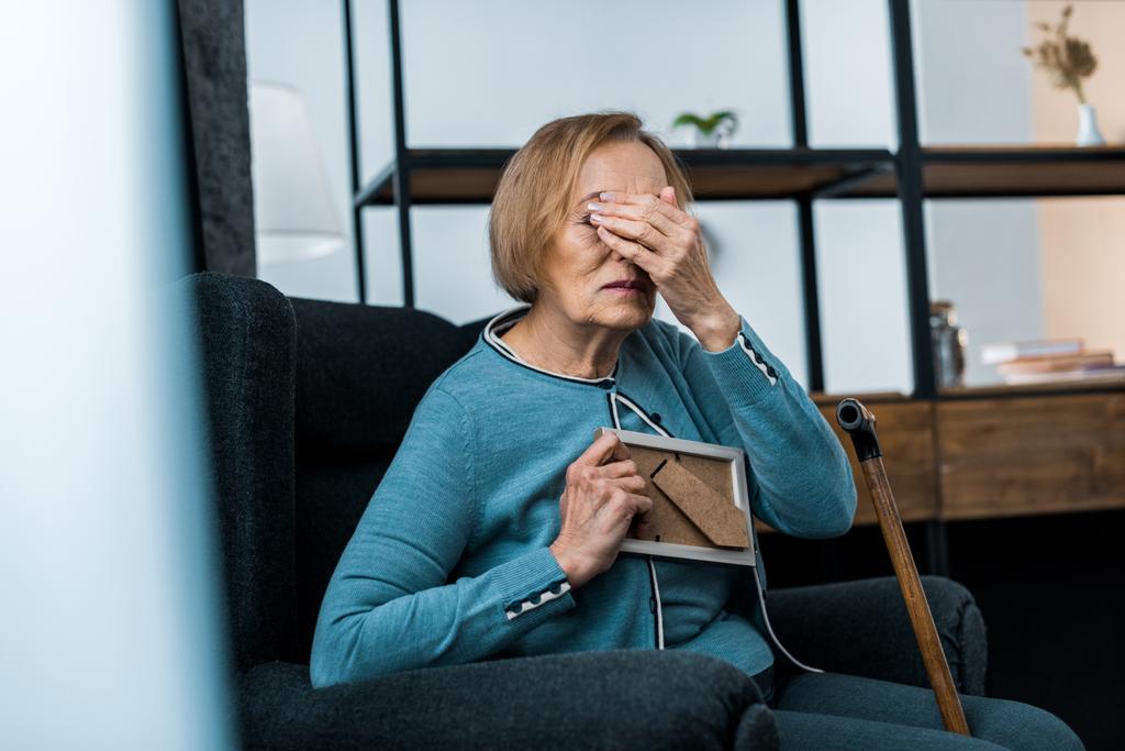 Rozpacza kobieta senior siedzący w fotelu, obejmujących twarz ręką i płacz trzymając ramki na zdjęcia - Zdjęcie, obraz