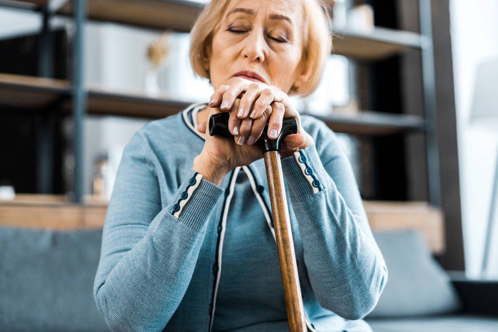 sad senior woman with eyes closed leaning on walking stick at home - Photo, Image