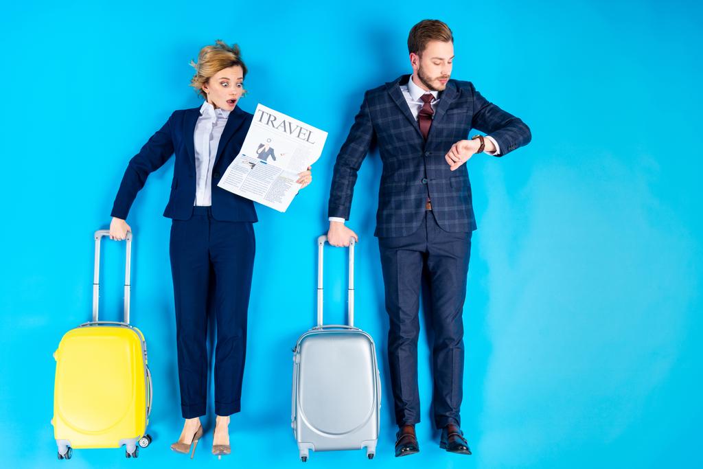 Surprised couple with suitcases looking at newspaper and watch on blue background - Photo, Image