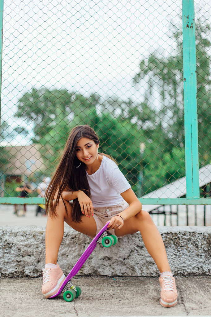 Zomer levensstijl beeld van trendy mooie jonge meisje zitten naast het skateboard coart met haar paarse plastic skateboard. - Foto, afbeelding