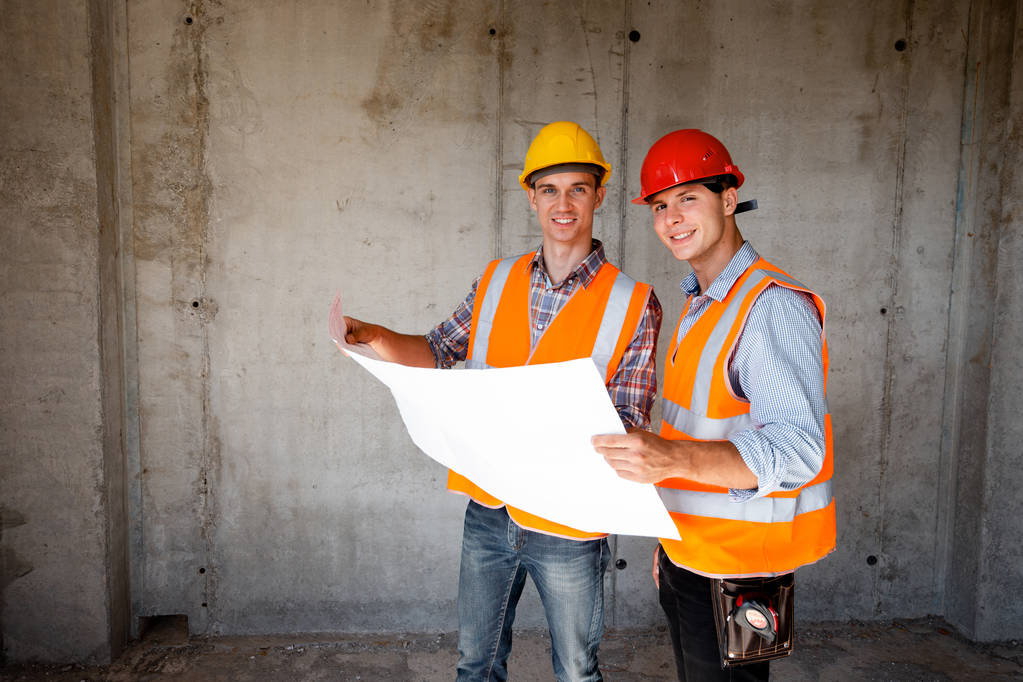 Twee mannen gekleed in het oranje werk vesten en helmen verkent bouw documentatie op een betonnen muur achtergrond - Foto, afbeelding