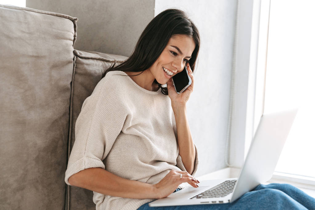 lächelnde junge Frau mit Laptop, während sie auf dem Boden auf einer Couch sitzt und mit dem Handy telefoniert - Foto, Bild