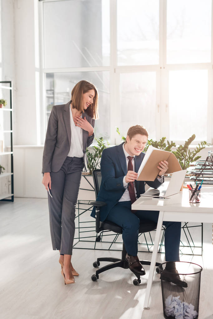привлекательная деловая женщина, стоящая со смартфоном рядом с счастливым коллегой и смеющаяся, глядя на документы в офисе
 - Фото, изображение