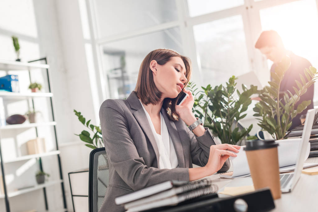 atractiva mujer de negocios hablando en el teléfono inteligente mientras está sentado en la oficina cerca de compañero de trabajo
 - Foto, Imagen
