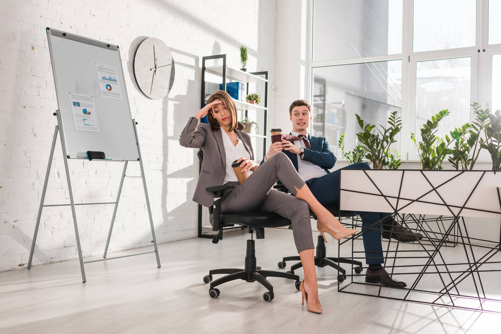 tired businesswoman holding paper cup and sitting near coworker in office  - Photo, Image