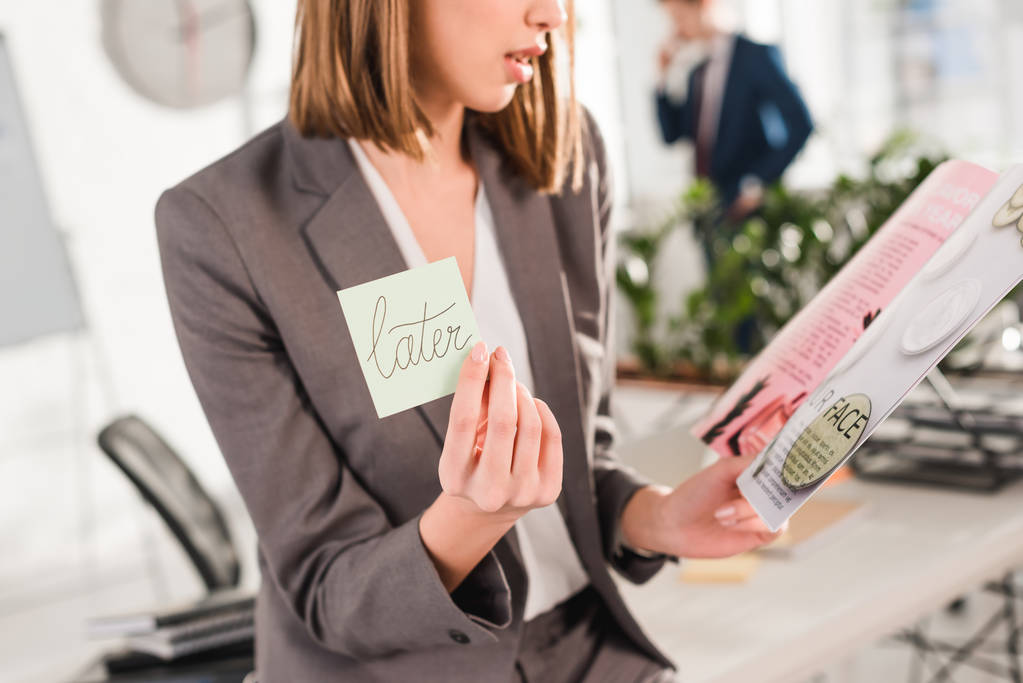 Ausgeschnittene Ansicht einer Geschäftsfrau mit klebrigem Zettel mit späterem Schriftzug und Magazin in der Hand mit Mitarbeiterin im Hintergrund, Verzögerungskonzept - Foto, Bild