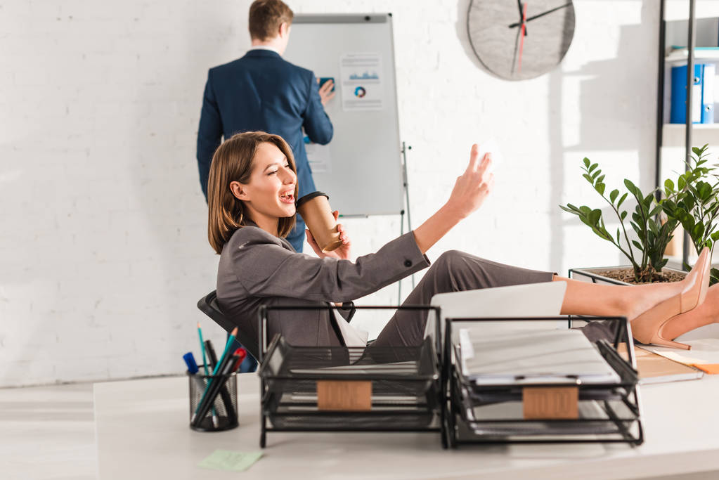 alegre empresaria tomando selfie con taza de papel cerca de compañero de trabajo en la oficina, concepto de dilación
 - Foto, Imagen