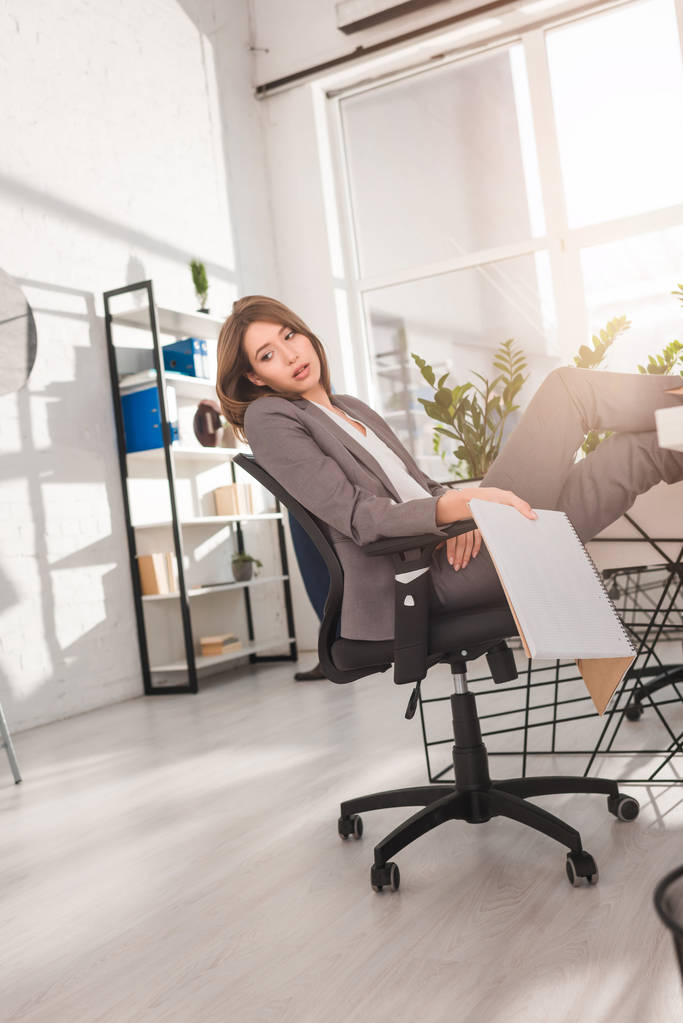  atraente empresária descansando na cadeira e segurando notebook com página em branco no escritório
 - Foto, Imagem