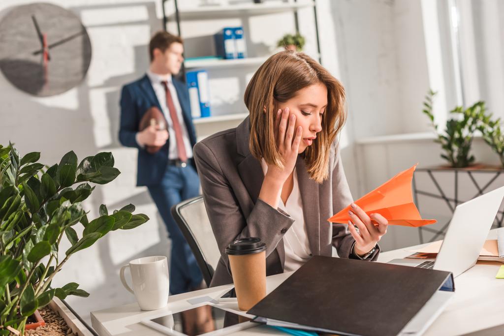 Müde Geschäftsfrau hält Papierflieger neben Laptop mit Mitarbeiterin im Hintergrund, Zauderkonzept - Foto, Bild