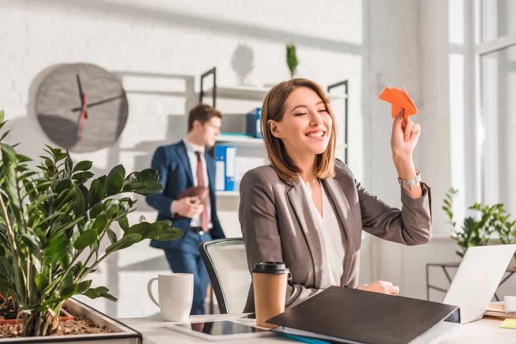 fröhliche Geschäftsfrau hält Papierflieger neben Laptop und Pappbecher mit Mitarbeiterin im Hintergrund, Zauderkonzept - Foto, Bild