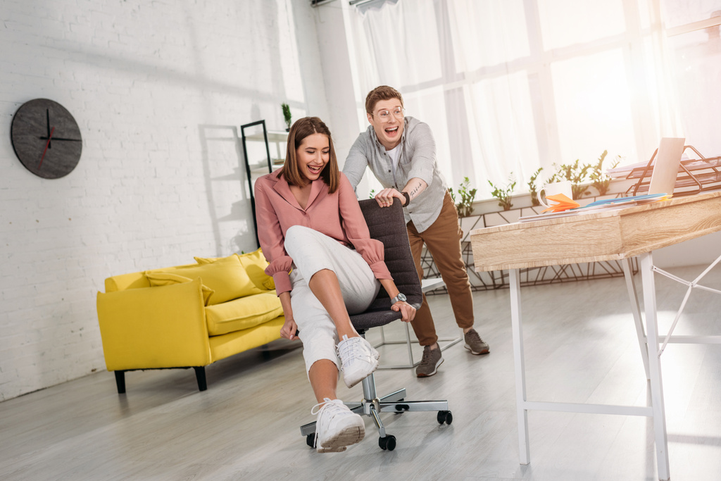 cheerful man smiling while pushing chair of happy girlfriend  - Photo, Image