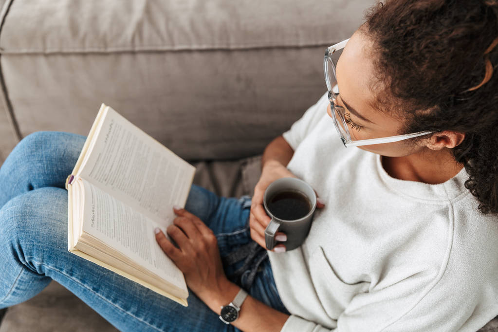 Image of brunette african american girl reading book and drinking tea while sitting on sofa in bright flat - Photo, Image
