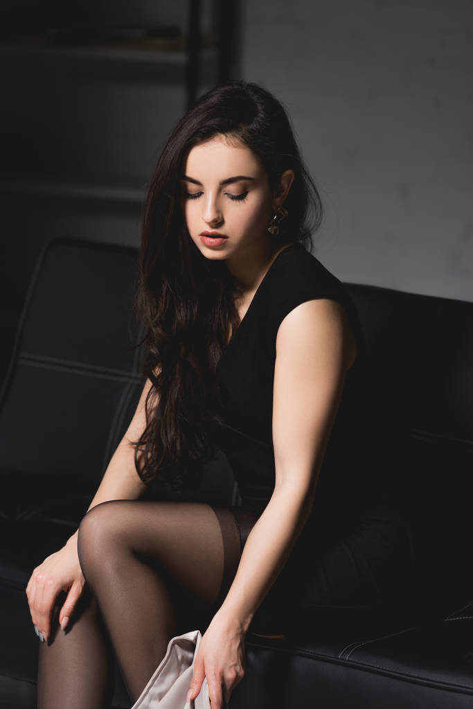 atraktivní brunetka žena v elegantních šatech sedící na gauči na tmavém pozadí - Fotografie, Obrázek