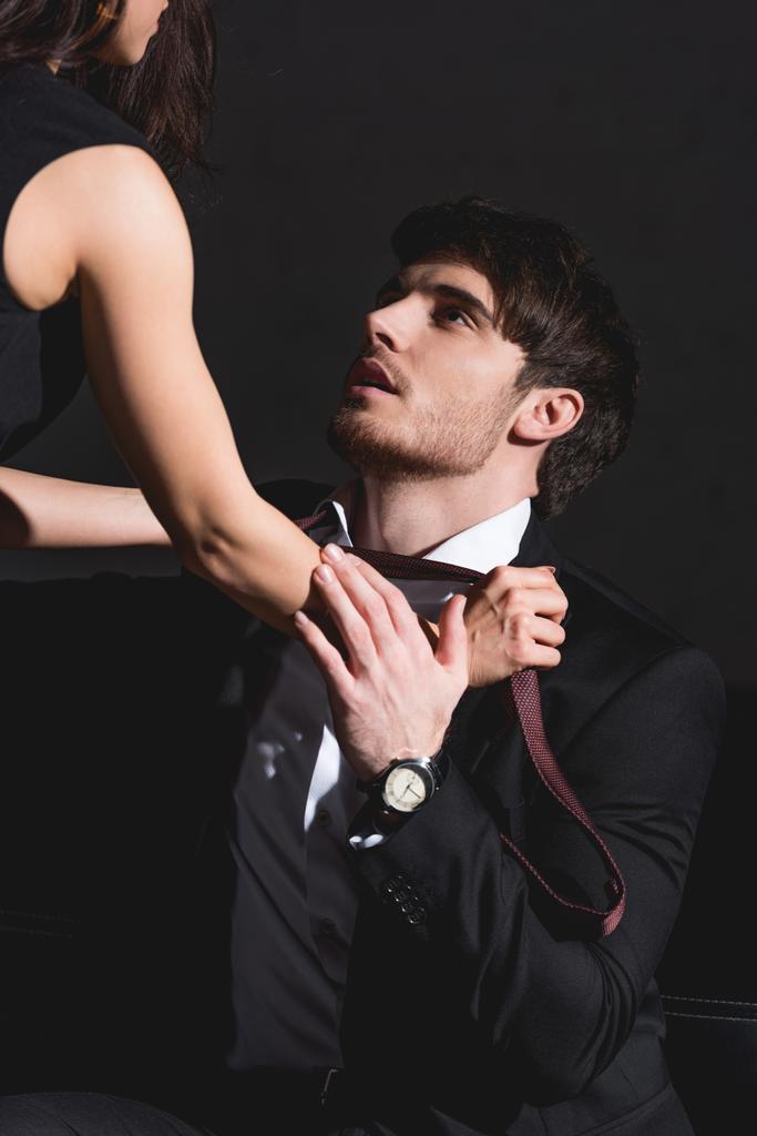 женщина в черном платье снимает галстук с красивым мужчиной в костюме, сидящим на диване на черном фоне
  - Фото, изображение