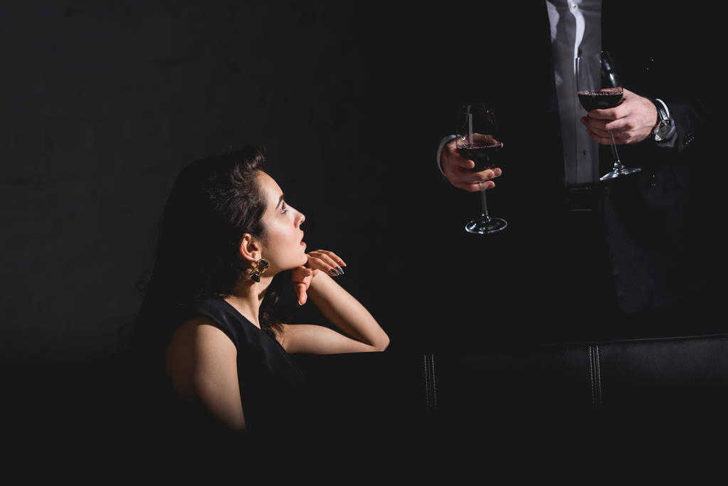 atractiva mujer sentada en vestido negro astuto hombre guapo en traje de pie detrás del sofá y la celebración de vasos con vino sobre fondo negro
 - Foto, imagen