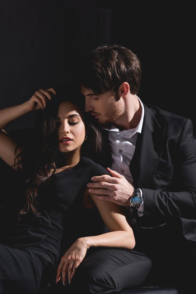 красивий чоловік у костюмі сидить на дивані, обіймає і цілує привабливу брюнетку жінку в чорному платті ізольовано на чорному
 - Фото, зображення