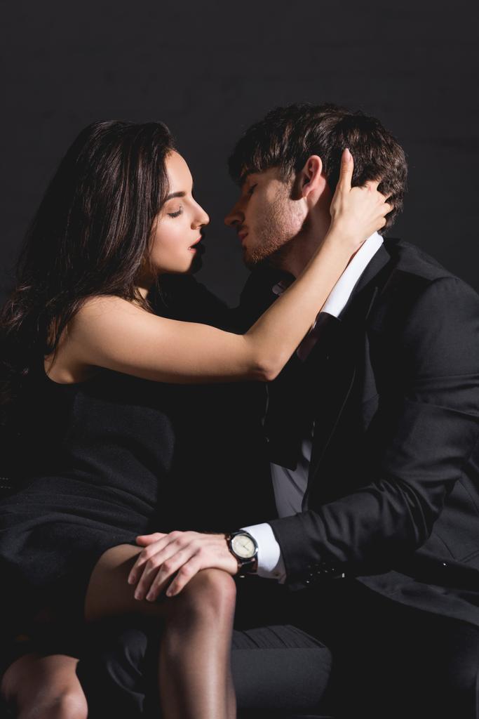 man in suit and woman in black dress sitting and kissing on couch on black background  - Photo, Image