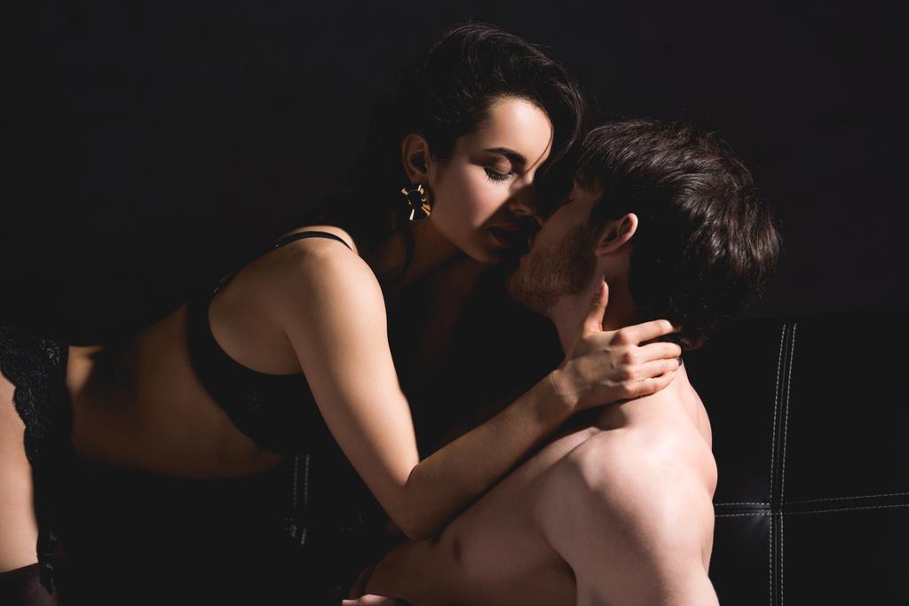 shirtless άνδρας και γυναίκα στο μαύρο εσώρουχα φιλιά στην απομονωμένη στο μαύρο καναπέ - Φωτογραφία, εικόνα