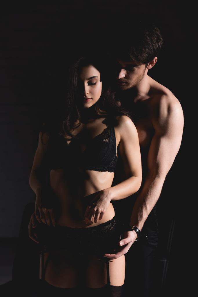 attraktive brünette Frau in schwarzen Dessous und hemdlos schöner Mann steht und umarmt isoliert auf schwarz - Foto, Bild