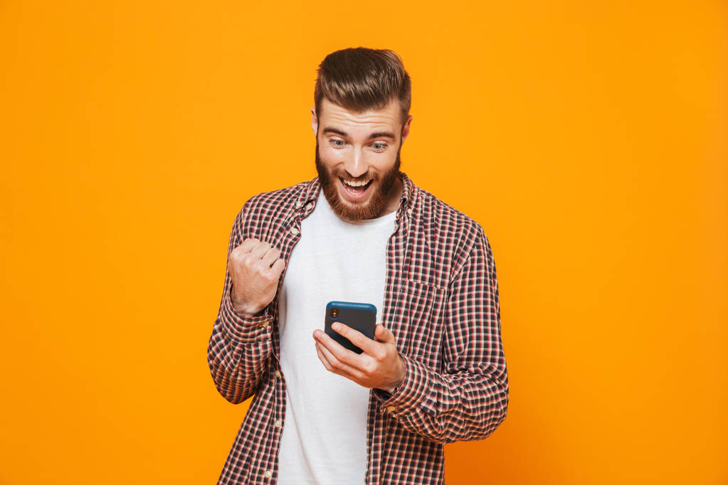 Porträt eines fröhlichen jungen Mannes in lässiger Kleidung, der isoliert vor gelbem Hintergrund steht und Mobiltelefone benutzt - Foto, Bild