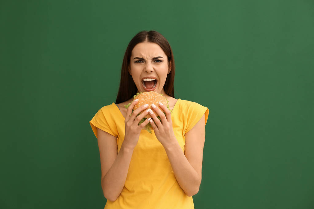 Genç kadın renk arka plan üzerinde lezzetli hamburger yemek - Fotoğraf, Görsel