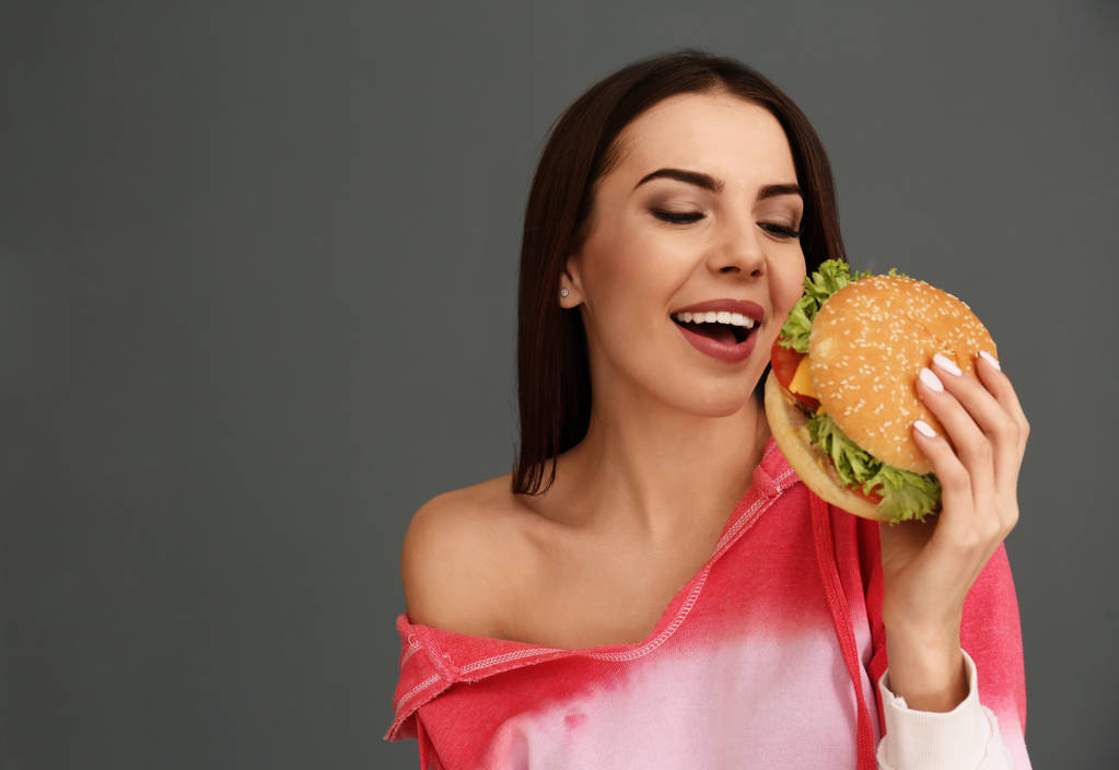 Молодая женщина ест вкусный бургер на сером фоне. Пространство для текста
 - Фото, изображение