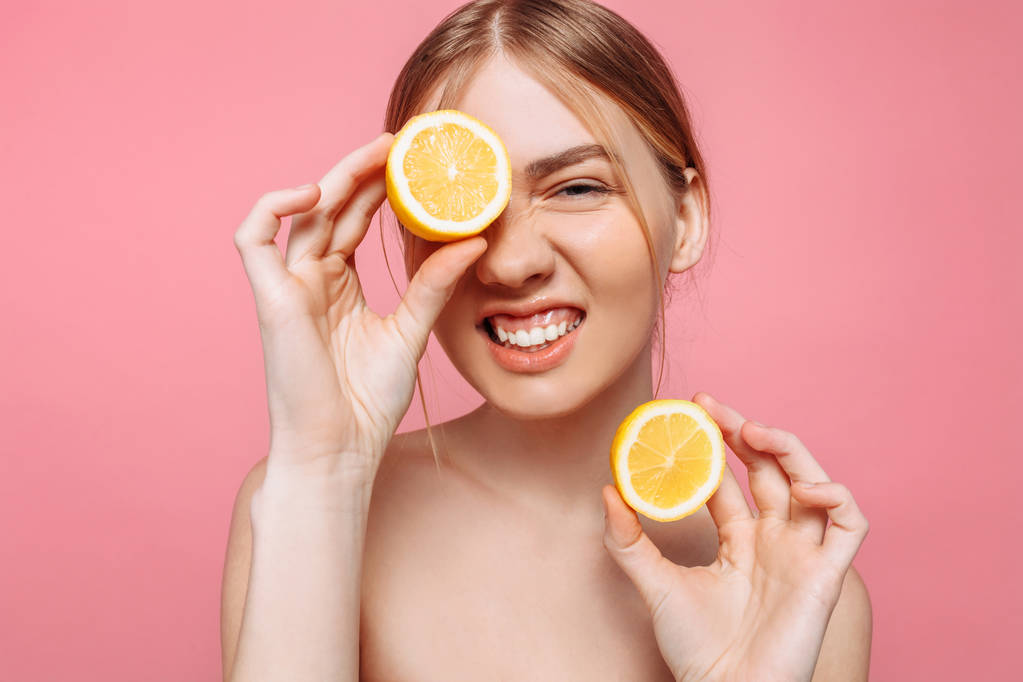 Retrato de una chica femenina alegre, piel natural y clara, una chica con dos trozos de limón, cubriendo un ojo con un limón, aislado sobre un fondo rosa. Concepto de cuidado de la piel, belleza natural
 - Foto, imagen